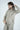 With medium-length hair, a person adjusts their Jaxson Maximus Deerfield Hoodie in Dark Khaki and matching pants. This light beige hoodie is crafted from super soft cotton and features a zip-up style against a plain, light-colored background.