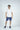 A man stands against a white background in a casual outfit featuring a Big League Notched V-Neck Short Sleeve shirt, blue shorts, and navy sneakers.