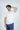 A man with short, wavy brown hair and a beard is wearing the Big League Notched V-Neck Short Sleeve White tee and blue pants. He stands against a light background, looking forward with one hand on the back of his head.