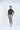 A man in the Sarasota Polo: Golfer With Palm Black and gray shorts stands against a white background. With his left hand in his pocket and white sneakers, his attire offers luxury performance fabric with four-way stretch for ultimate comfort.