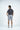 A man stands backwards in a light gray studio, highlighting the Sarasota Polo: Golfer With Palm Black, crafted from luxurious performance fabric. Hes wearing light gray shorts with four-way stretch and black and white sneakers for effortless style and comfort.