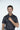 A man with short dark hair stands against a plain background, wearing the Sarasota Polo: Golfer With Palm Black. This shirt features a luxury performance fabric with small white and pink designs. Its four-way stretch offers comfort and style as he thoughtfully holds the collar.
