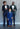 A young boy in a blue suit stands smiling between two adults in suits, one in navy and one in dark green. Both adults are pointing at the boy, also smiling. They are posing against a gray wall with visible bolts.