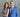 A woman with long brown hair in a sleeveless gray dress stands smiling with arms crossed next to a man in a blue suit and tie. Both are smiling and standing against a gray, textured wall.