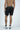 A shirtless man stands facing away, wearing a Jaxson Maximus Beach To Trail Short in Black with Gray. The short is made from quick-dry fabric.