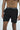 A shirtless person stands against a plain background, wearing Beach To Trail Short Black with Gray. These shorts, featuring quick-dry fabric, a drawstring, and a subtle logo near the hem, are perfect for versatile adventures.