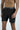 A shirtless person stands against a plain background wearing Beach To Trail Short Black with Gray. Their hands are by their sides, and the quick-dry fabric shorts feature a small logo on the left leg and include a drawstring.