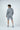 A brown-haired man stands facing away in a Jaxson Maximus Deerfield Hoodie in Charcoal Gray with matching Jaxson Maximus Deerfield shorts. He rests one hand in his pocket, in a well-lit studio, sporting gray sneakers with white soles made from super-soft cotton.