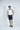 A dark-haired man with a beard stands against a white background, wearing the Sanibel Polo in White with Gray, made from luxury performance fabric. He pairs it with navy shorts and shoes, one hand in his pocket as he looks directly at the camera.