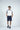 Against a light gray background, a man wears the Sanibel Polo in white with gray luxury performance fabric, navy shorts, a dark belt, and dark sneakers. With one hand in his pocket, he gazes to the side with a relaxed expression.