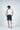 A man stands with his back turned, wearing the Sanibel Polo in white with gray, crafted from luxury performance fabric, paired with navy shorts and sneakers. Against a plain white backdrop, his ensemble ensures style and comfort through its four-way stretch.