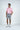 A man stands against a plain background, wearing a Dolphin Tail Pink West Palm Polo made from luxury performance fabric, gray shorts, and white athletic shoes. His hands rest at his sides as he gazes slightly right, embodying effortless style and comfort.