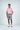 A man confidently wears a Dolphin Tail Pink West Palm Polo paired with gray shorts, white sneakers, and a patterned belt. His hands are relaxed, one by his side and the other in his pocket, set against a plain white backdrop.