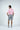 A man stands confidently in the Dolphin Tail Pink West Palm Polo made from luxury performance fabric, paired with gray shorts and white sneakers. His short dark hair contrasts with a plain white backdrop as he faces away, hands on hips.