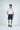 A man in a West Palm Polo, White With Gray Stripes, stands against a plain background. Sporting navy shorts, sneakers, short hair, and a beard, he gazes left with relaxed hands by his sides in an effortlessly stylish look.