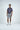 A man in the Sarasota Polo: Golf Girl Navy, crafted from luxury performance fabric, and gray shorts stands against a white background. With white sneakers and short dark hair, he exudes casual elegance. His hands are relaxed by his sides as he gazes slightly to the left.
