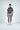 A man stands against a plain white background, showcasing the Sarasota Polo: Golf Girl Navy made of luxury performance fabric. He pairs it with gray shorts, white sneakers, and a brown belt. His relaxed hands by his sides highlight the comfort and style of his outfit.
