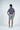 A person wearing the Sarasota Polo: Golf Girl Navy, featuring a navy patterned short-sleeve design in luxury performance fabric, paired with gray shorts and white sneakers is standing with their back turned on a plain white background.