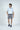 Wearing the West Palm Polo in Gingham Light Blue, paired with gray shorts and dark sneakers, a person stands against a plain backdrop. With one hand in their pocket, they glance to the side.
