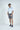 A man with short, curly hair and a beard stands against a white background, wearing a Gingham Light Blue West Palm Polo made from luxury performance fabric, gray shorts, and navy sneakers. He looks to his left with his hands in his pockets.