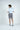 A brown-haired man stands against a white background, wearing the West Palm Polo: Gingham Light Blue made of luxury performance fabric, paired with gray shorts and sneakers. His hands rest near his waist as he gazes to the side, slightly turned away from the camera.