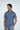 A man stands against a light gray background wearing the Panhandle Polo: Tropical Oasis, a navy blue wrinkle-resistant short-sleeve shirt with a white leaf pattern and breathable Micro-Pique fabric. He gazes left, one hand in his light-colored pants pocket.