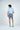 A man with brown hair, dressed in a light blue and white striped West Palm Polo made from luxury performance fabric and gray shorts, stands with his back to the camera. He is in a neutral pose against a plain white background, glancing to his right.