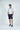 A man with short hair stands against a plain white background, wearing a West Palm Polo: Gingham Gray. His light polo shirt and dark shorts provide four-way stretch for comfort, with his hands in pockets exuding a relaxed vibe.