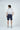 A man in the West Palm Polo: Gingham Gray shirt, navy shorts, and gray sneakers stands with his short brown hair contrasting a plain gray background, illustrating luxury performance fabric.