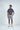 A man stands against a white background in the West Palm Polo: Navy with White Stripes, crafted from luxury performance fabric. He pairs it with light gray shorts and white athletic shoes. His hands in his pockets, he smiles softly, gazing to the side.