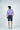 Man stands against a light gray background, facing away. Hes sporting the Sarasota Polo: Golf Cart Palm Purple, made from luxury performance fabric. The ensemble is completed with navy shorts and gray sneakers.