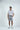 A man in the West Palm Polo: White With Navy Stripes and gray shorts stands against a plain white background. He has one hand in his pocket, smiles, wears sneakers, and pairs it with a dark belt.