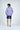 A person with wavy brown hair stands facing away, wearing a Key West 1/4 Zip: Golf Cart Palm shirt in purple patterns available in multiple sizes, navy shorts, and gray sneakers with blue accents. The serene backdrop is light gray.