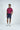 A man stands against a plain backdrop, wearing a Sarasota Polo: Golf Girl Ruby crafted from luxury performance fabric. He pairs it with dark blue shorts and white sneakers. With one hand in his pocket and the other by his side, he gazes to the side with a neutral expression.