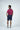 A man with dark hair stands with his back to the camera, wearing a Sarasota Polo: Golf Girl Ruby short-sleeve shirt made of luxury performance fabric, dark blue shorts, and white shoes. The plain background subtly contrasts with his casual yet refined look.