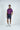 A man stands against a plain white background wearing the Sarasota Polo: Shark Attack Purple, made from luxury performance fabric. Paired with navy shorts and white sneakers, exuding effortless style, he poses confidently with one hand in his pocket and a slight smile.