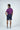 A man faces slightly left, hands in pockets, wearing a Sarasota Polo: Shark Attack Purple made from luxury performance fabric. Paired with navy shorts and white sneakers, the polos four-way stretch promises comfort and style against a plain background.
