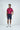A man smiles in a Sarasota Polo: Shark Attack Ruby made from luxury performance fabric, paired with dark blue shorts and white sneakers. One hand is in his pocket, the other by his side, set against a plain white background that highlights the polos elegance.