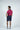 A man in a Sarasota Polo: Shark Attack Ruby and navy shorts stands with his back to the camera, showcasing the luxury fabric. Hands in pockets, white sneakers complete his stylish look. The plain background accentuates his four-way stretch appeal.