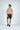 A shirtless man stands smiling and looking left, wearing gray athletic shorts with a sky-blue drawstring made from quick-dry fabric. His look is completed with black athletic shoes featuring white soles, perfect for a beach-to-trail transition against a plain white backdrop.