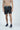 A shirtless person stands against a white background, wearing Beach To Trail gray shorts with a light blue drawstring and navy blue sneakers.