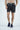 A person wearing Beach To Trail Short Gray with Sky Blue, made from quick-dry fabric, stands against a plain background. Facing forward and with hands in pockets, they are ready for any adventure while sporting their blue sneakers.